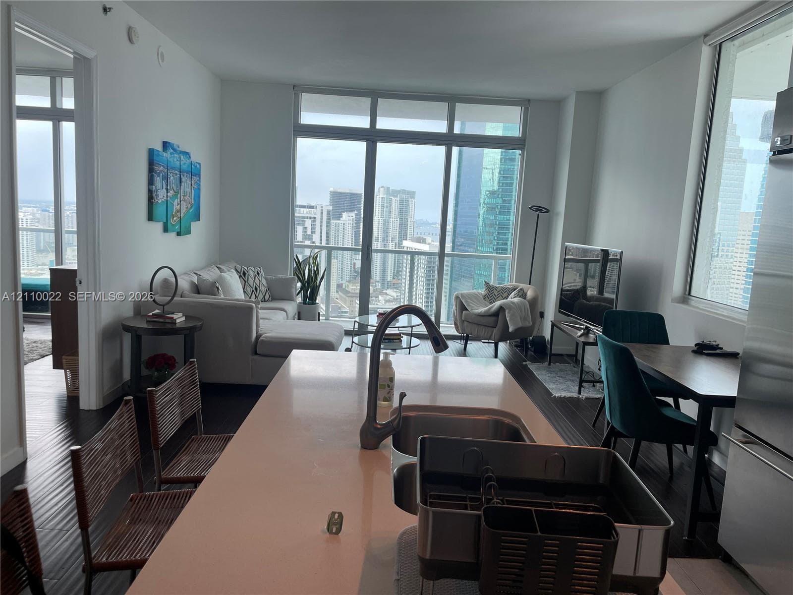 Open-concept interior featuring wood-finish flooring, a kitchen island with a neutral solid-surface countertop, an undermount stainless steel sink, and floor-to-ceiling windows
