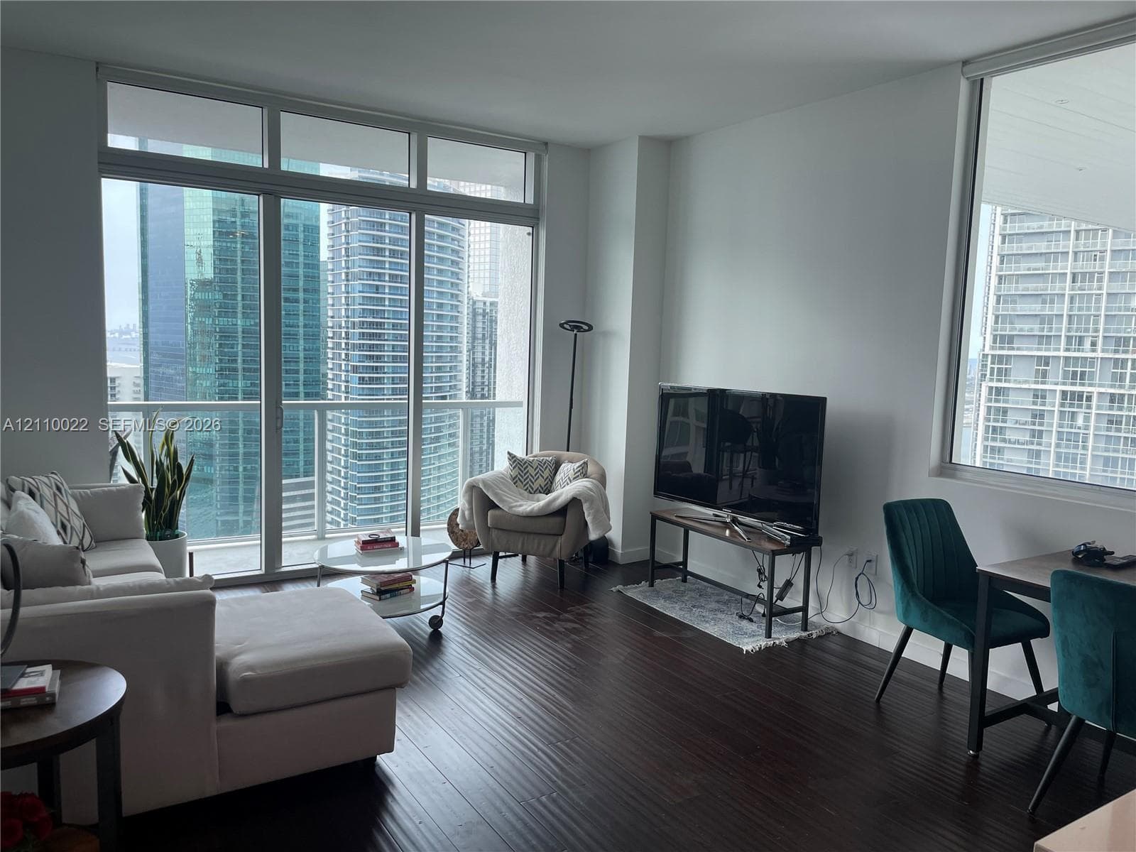 Floor-to-ceiling windows, dark wood-finish flooring, glass-paneled balcony access, and high-rise urban scenery