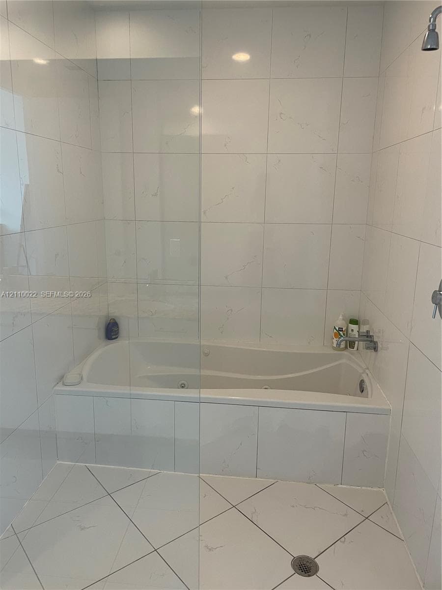 Built-in jetted bathtub featuring large-format light-toned tile surround, glass partition, and diamond-patterned tile flooring