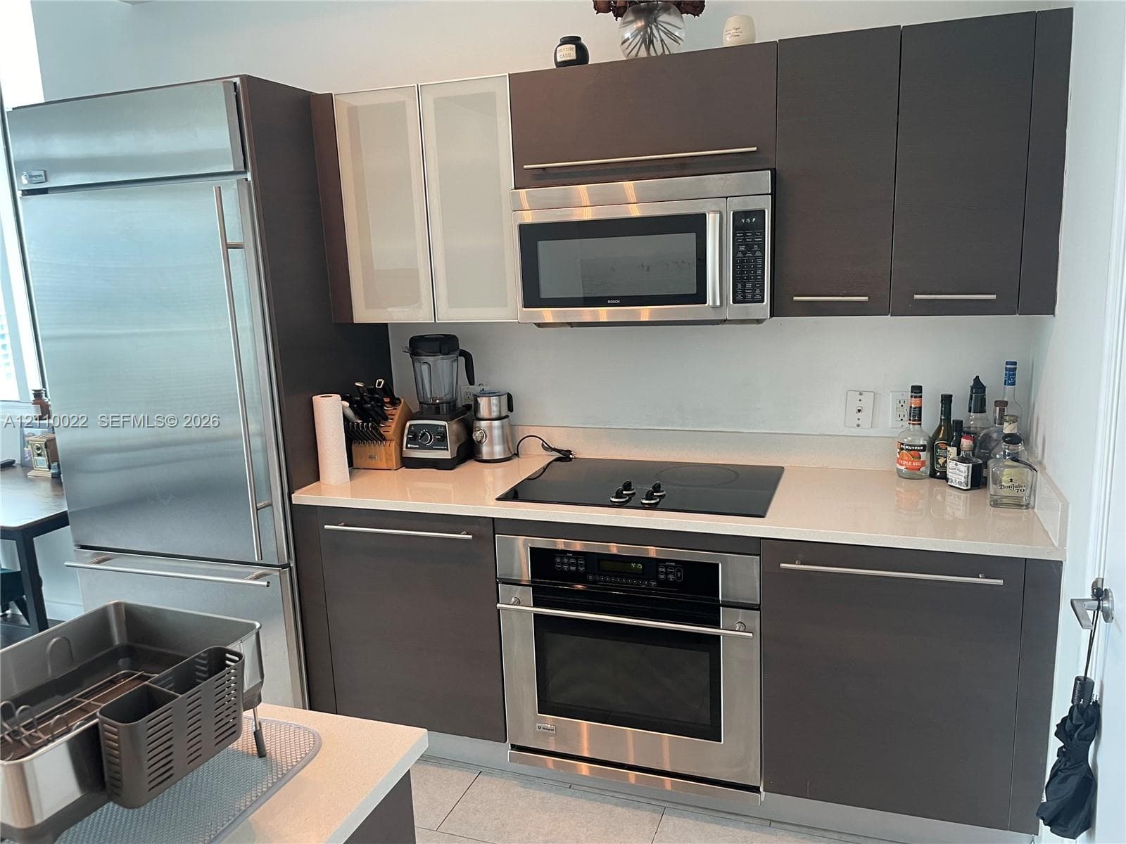 Kitchen featuring flat-panel dark wood-finish cabinetry, light-toned solid surface countertops, a built-in stainless steel refrigerator, an integrated glass-top cooktop, and a built-in stainless steel wall oven