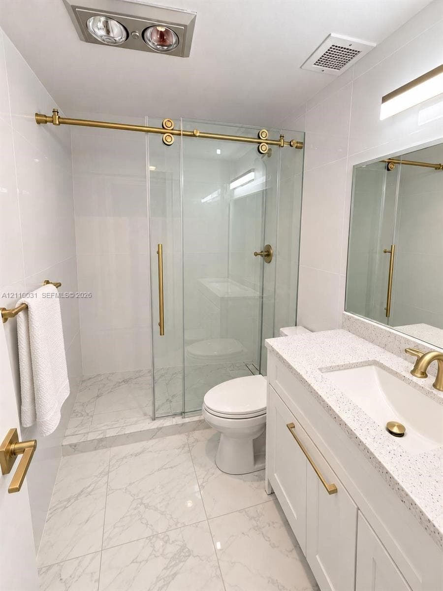 Bathroom featuring a walk-in shower with a glass sliding door, brass hardware, stone-finish tile flooring, quartz-surface vanity countertop, and white shaker-style cabinetry