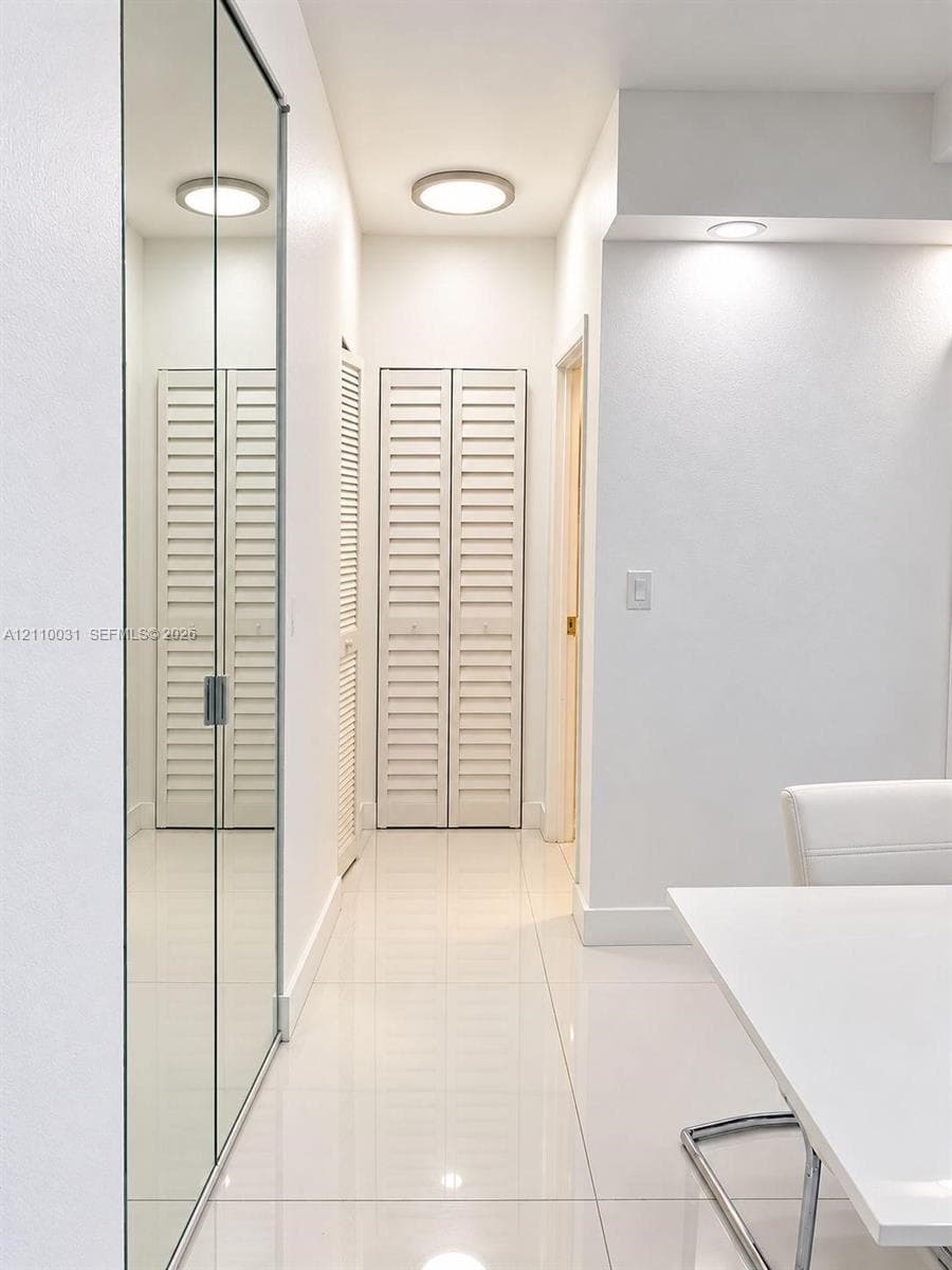 Hallway featuring floor-to-ceiling mirrored sliding closet doors, white louvered bifold closet doors, high-gloss tile flooring, and flush-mount ceiling lighting