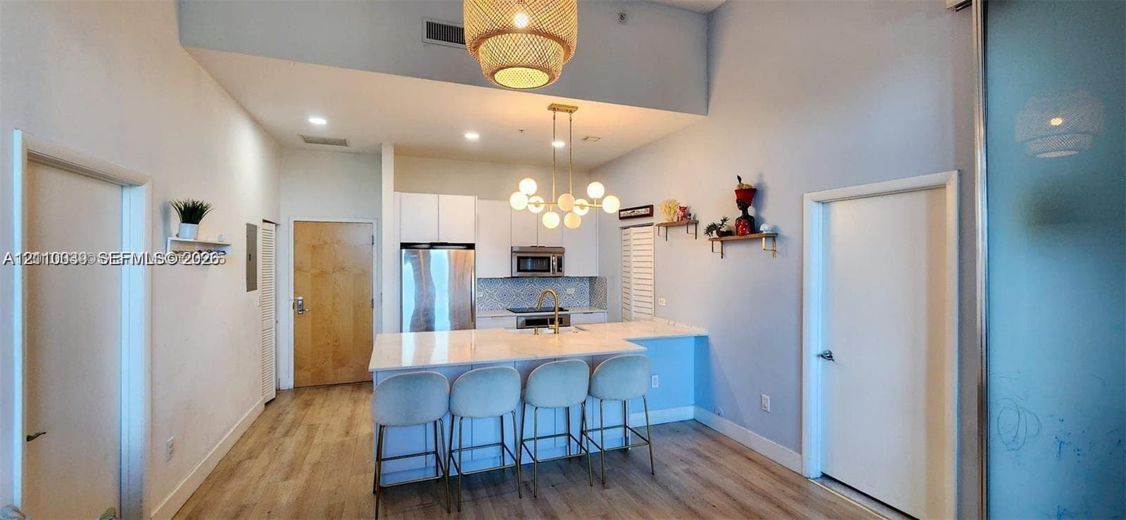 Modern kitchen featuring white flat-panel cabinetry, light-toned stone-surface island, stainless steel appliances, tiered globe chandelier, and light wood-finish flooring