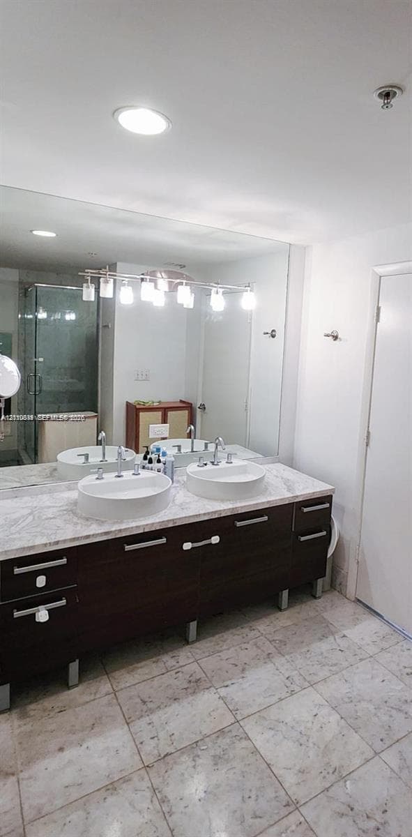 Double vanity featuring dark wood-finish cabinetry, a stone-style countertop, dual round drop-in sinks, and a large-format mirror with integrated overhead lighting