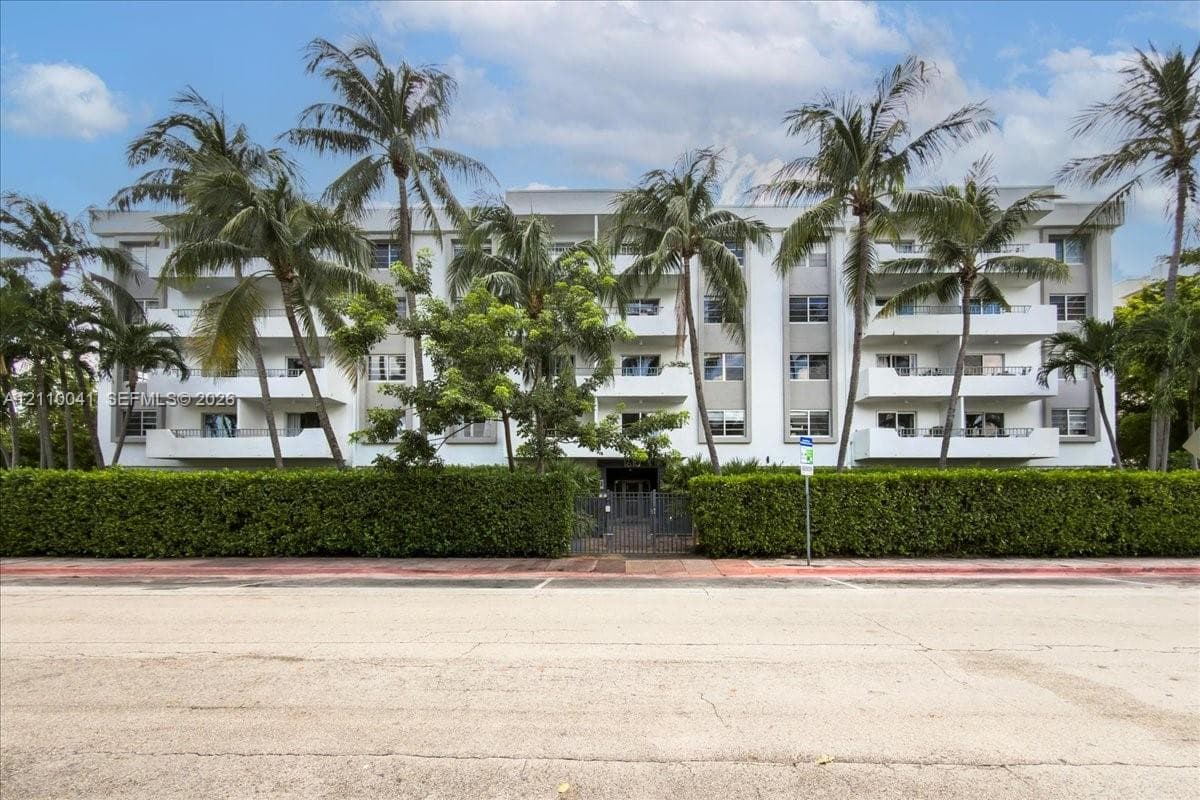 Multi-story residential building featuring private balconies, vertical architectural pillars, and mature palm tree landscaping set behind a manicured hedge