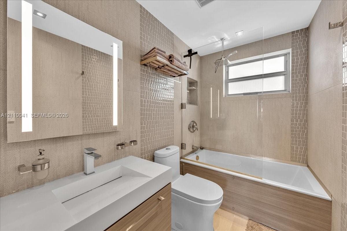 Bathroom featuring a wood-finish vanity with a white rectangular vessel sink, a wall-mounted LED-backlit mirror, a glass-enclosed shower and tub combination, and neutral textured tile wall accents