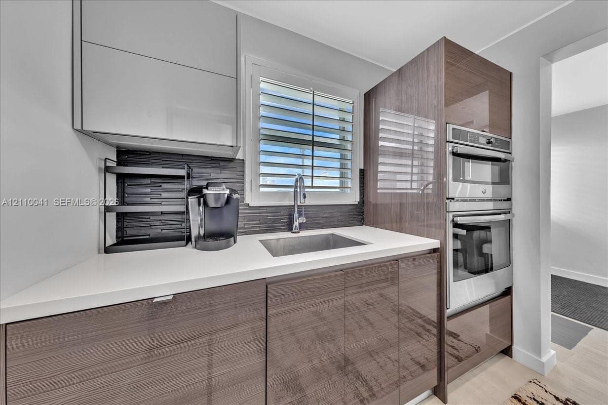 High-gloss cabinetry, stone-finish countertops, stainless steel dual wall oven, dark linear tile backsplash, and undermount stainless steel sink