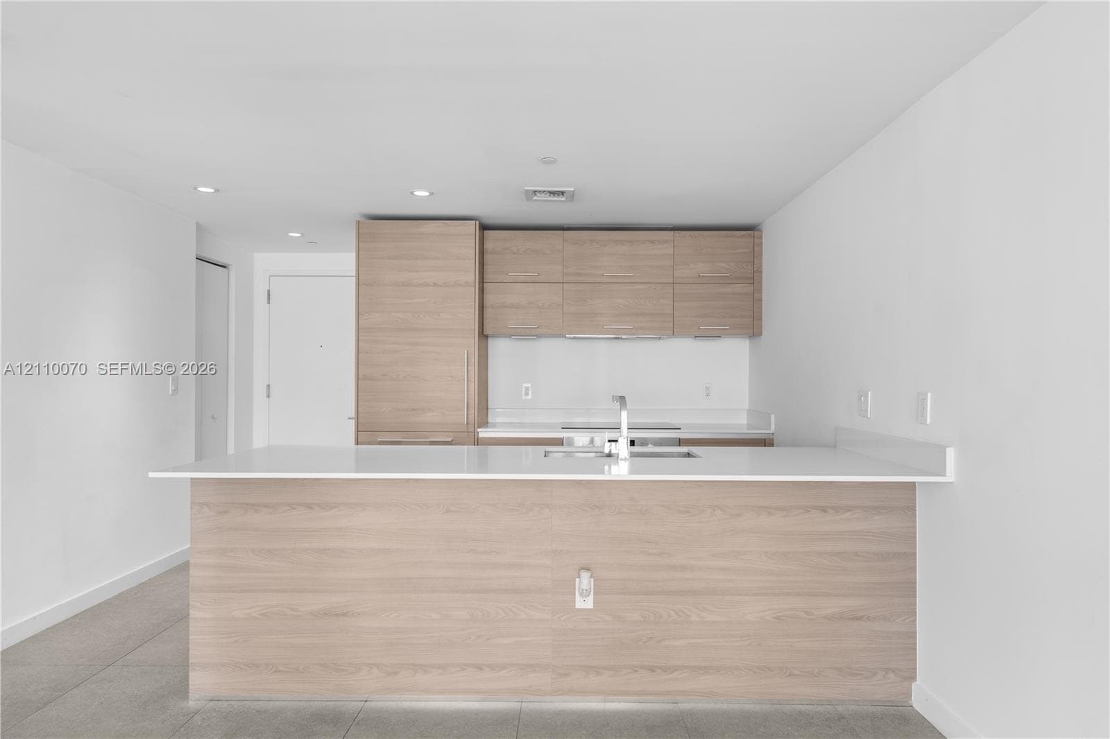 Kitchen featuring light wood-finish cabinetry, white solid-surface countertops, a center island with an integrated sink, and recessed ceiling lighting