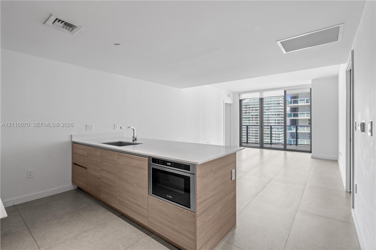 Kitchen island featuring wood-finish cabinetry, solid surface countertop, stainless steel sink with high-arc faucet, and built-in microwave, complemented by large-format tile flooring and floor-to-ceiling glass doors