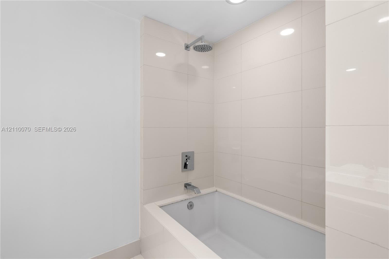 Full-height neutral wall tiling, integrated bathtub, chrome shower head, chrome wall-mounted faucet, and recessed ceiling lighting