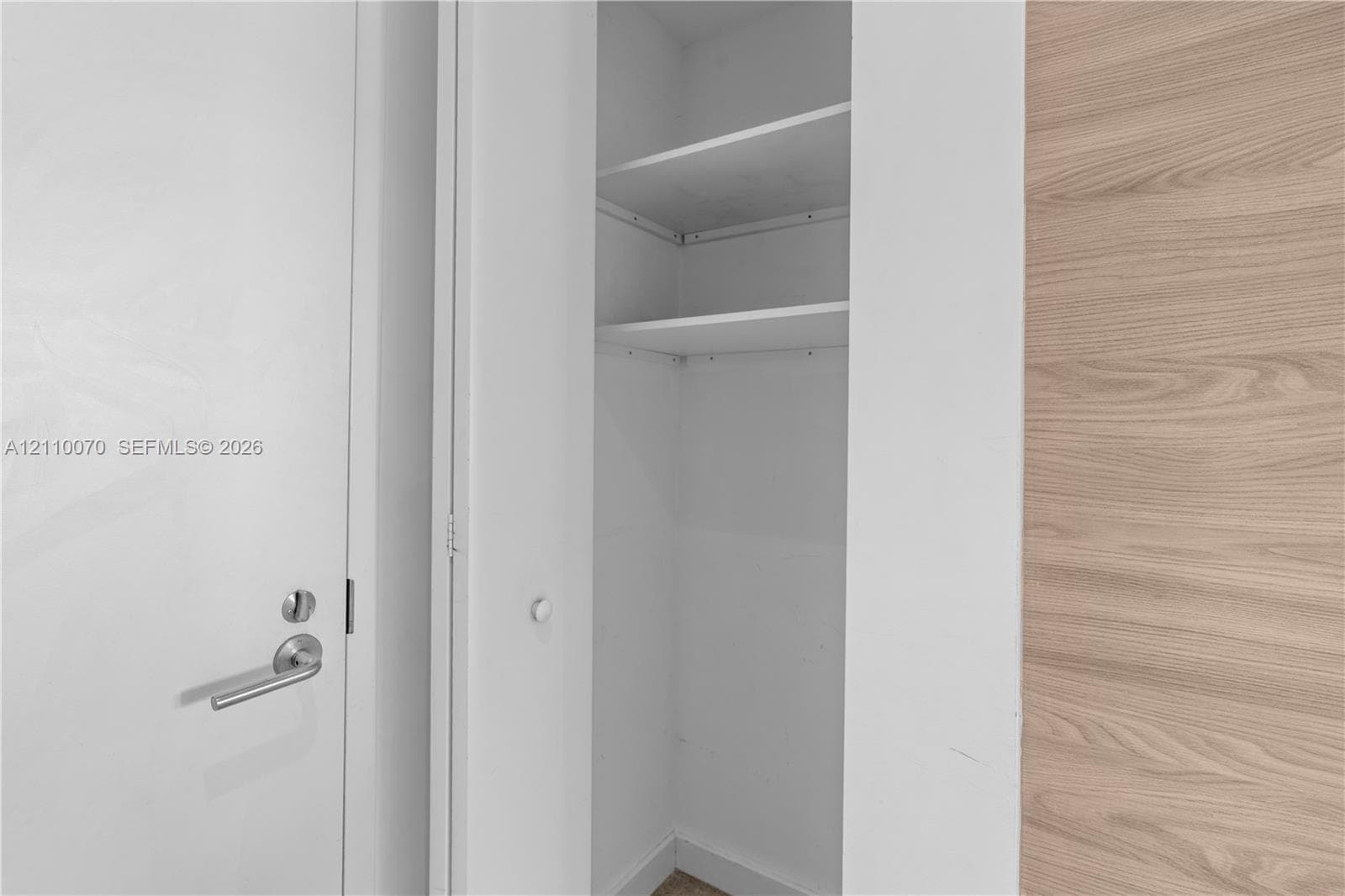 Built-in closet featuring white shelving, a solid white door with a satin-nickel lever handle, and a wood-finish wall panel