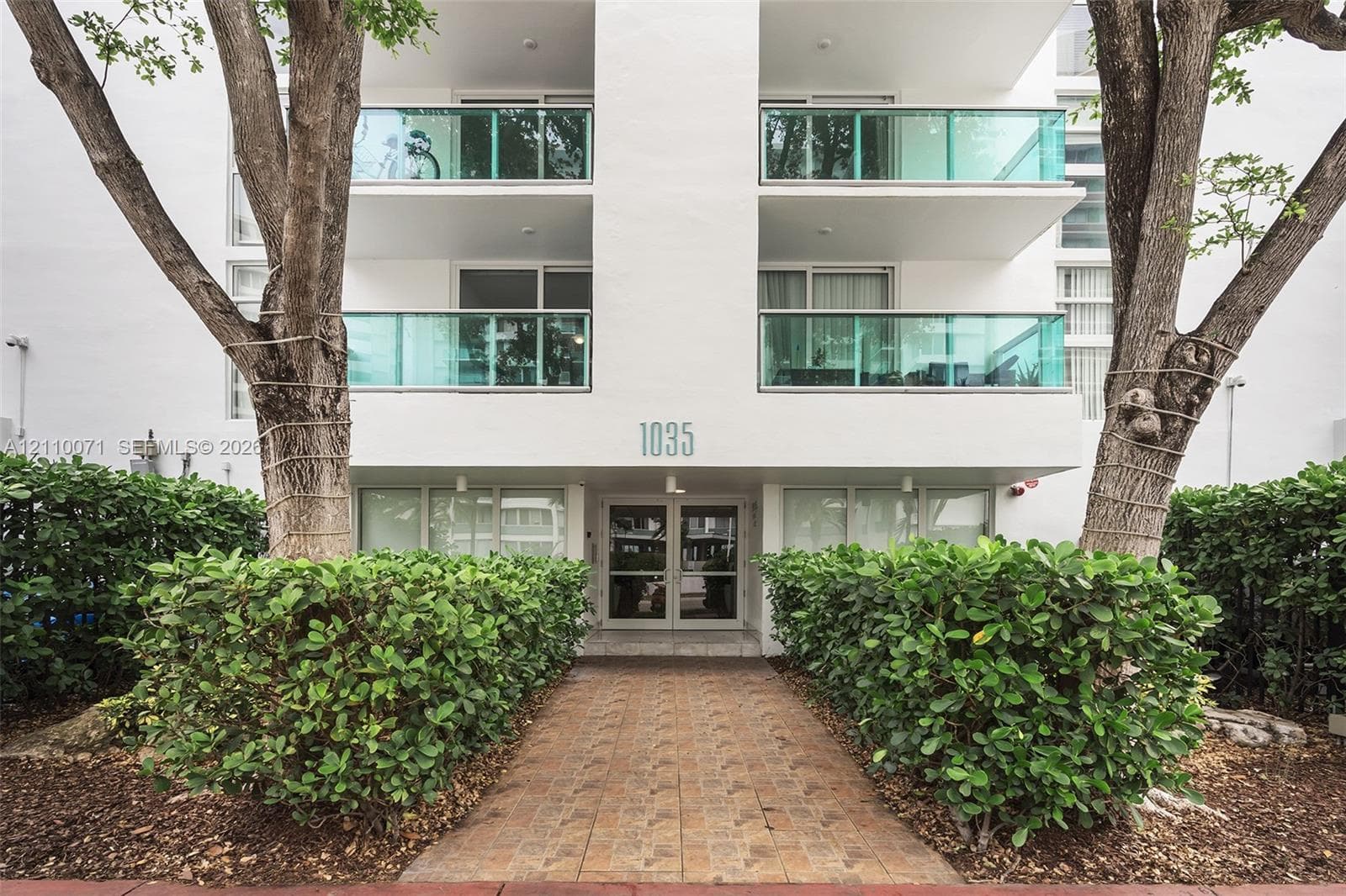 Exterior facade featuring white stucco finish, glass-railed balconies, a paved entryway, and mature landscaping