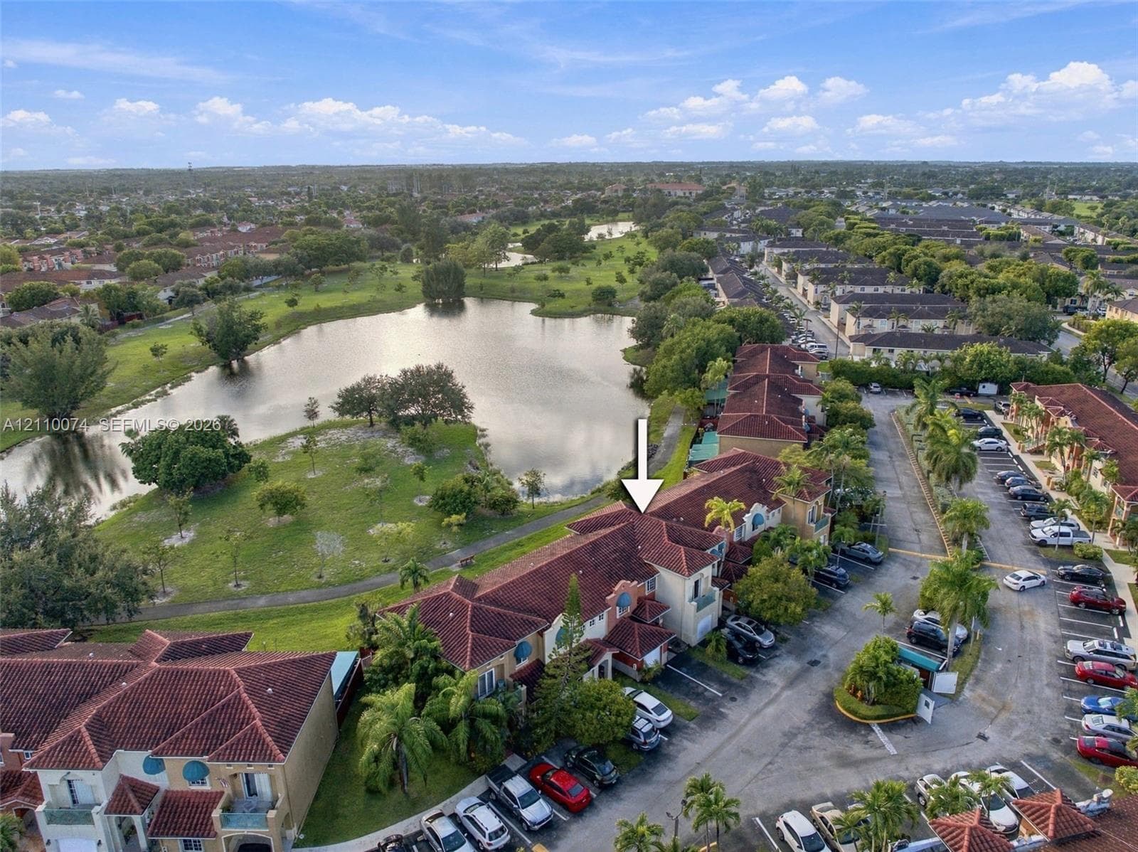 Aerial view featuring a multi-story residence with a multi-gabled tile roof, concrete exterior, balcony, and proximity to a community lake and green space