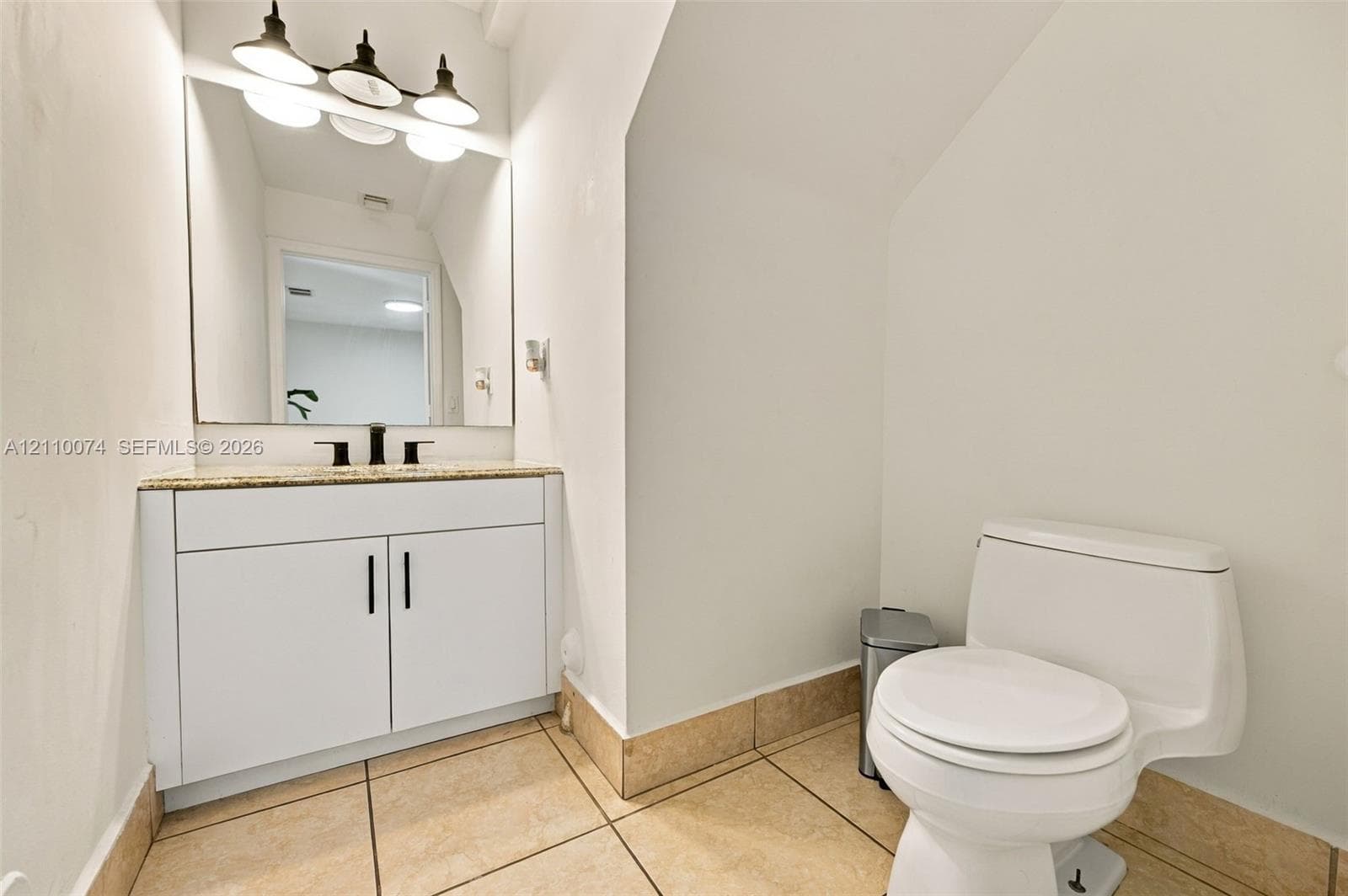 White vanity featuring stone-finish countertop, undermount sink with dark metal faucet, expansive wall mirror, three-light fixture, and ceramic tile flooring