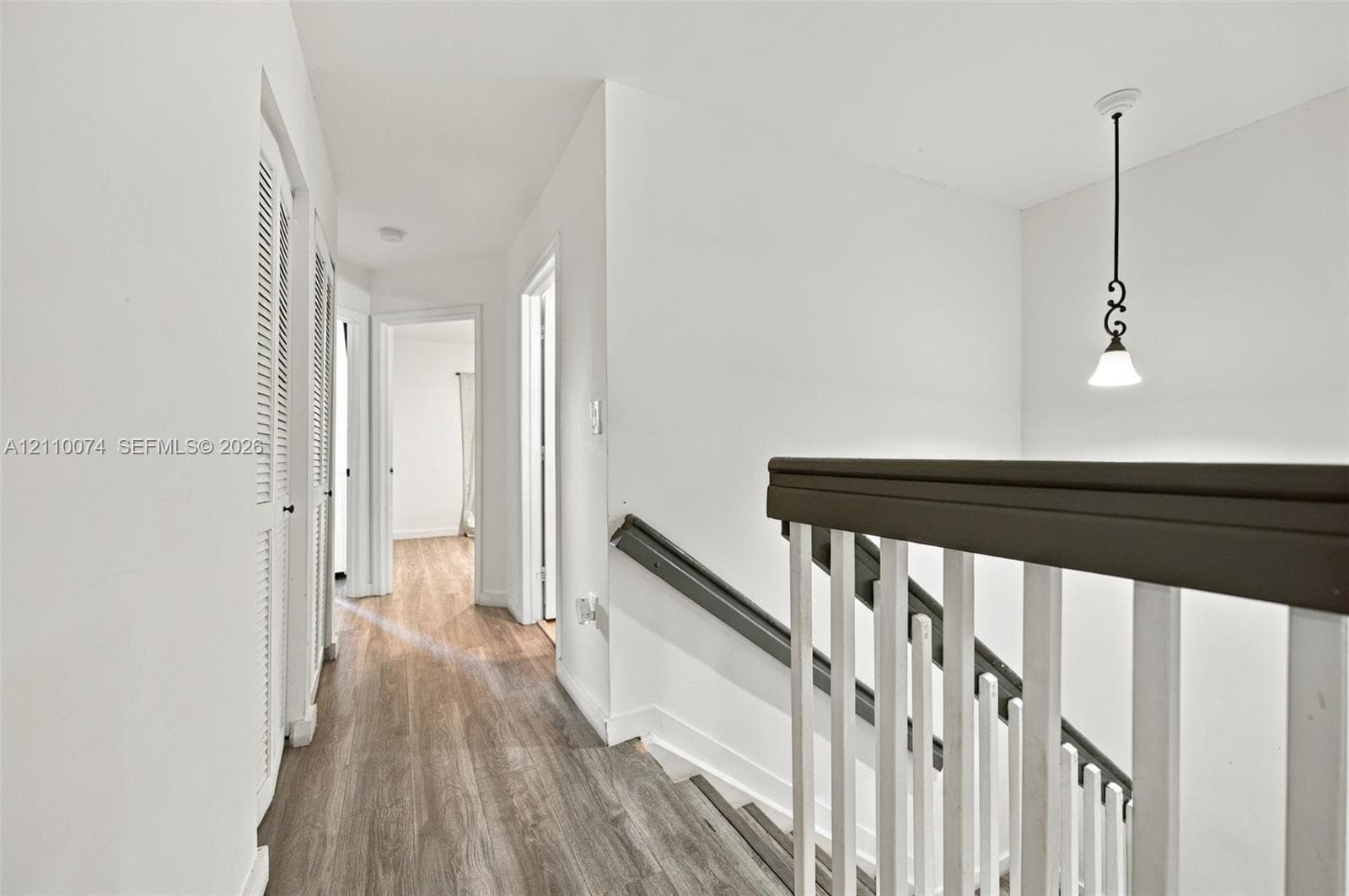 Hallway featuring wood-finish flooring, white louvered closet doors, and a staircase with a dark-finish handrail and white balusters, accented by a decorative hanging pendant light
