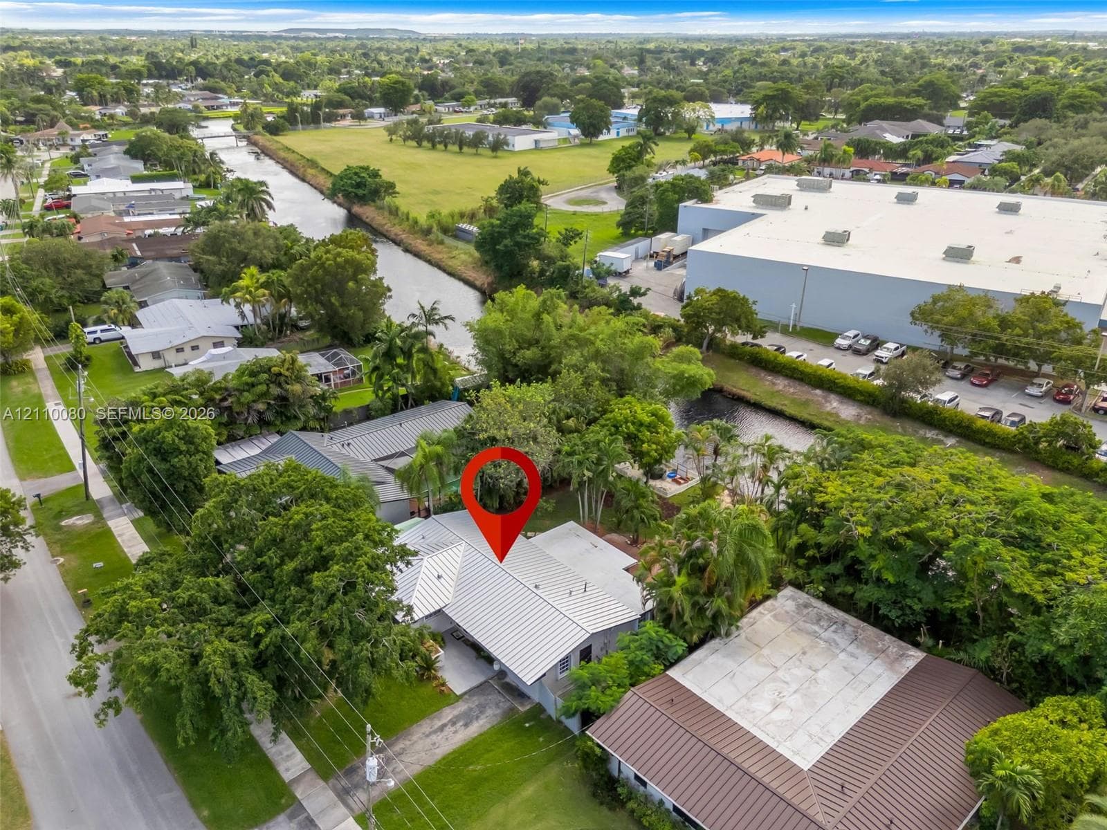 Aerial perspective featuring a metal-roofed residence, asphalt driveway, manicured landscaping, and mature trees situated on a canal-front lot