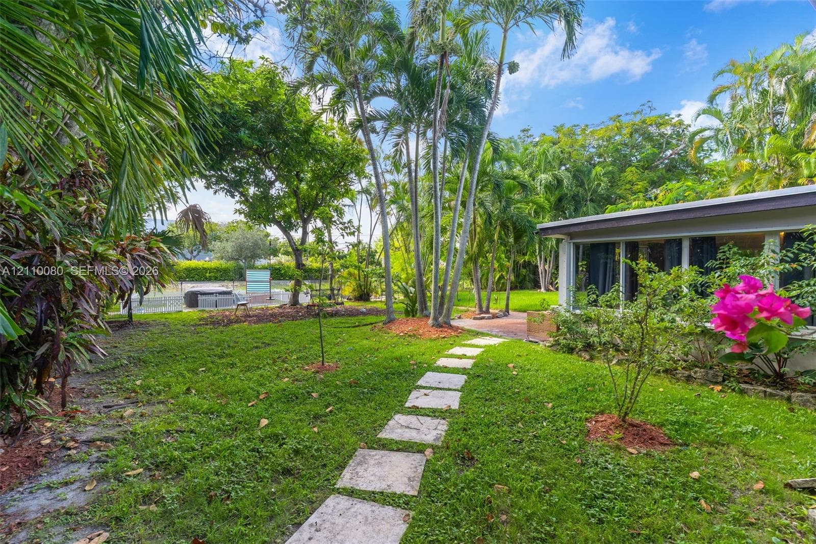 Landscaped backyard featuring mature palm trees, a stone paver walkway, brick patio, and lush vegetation