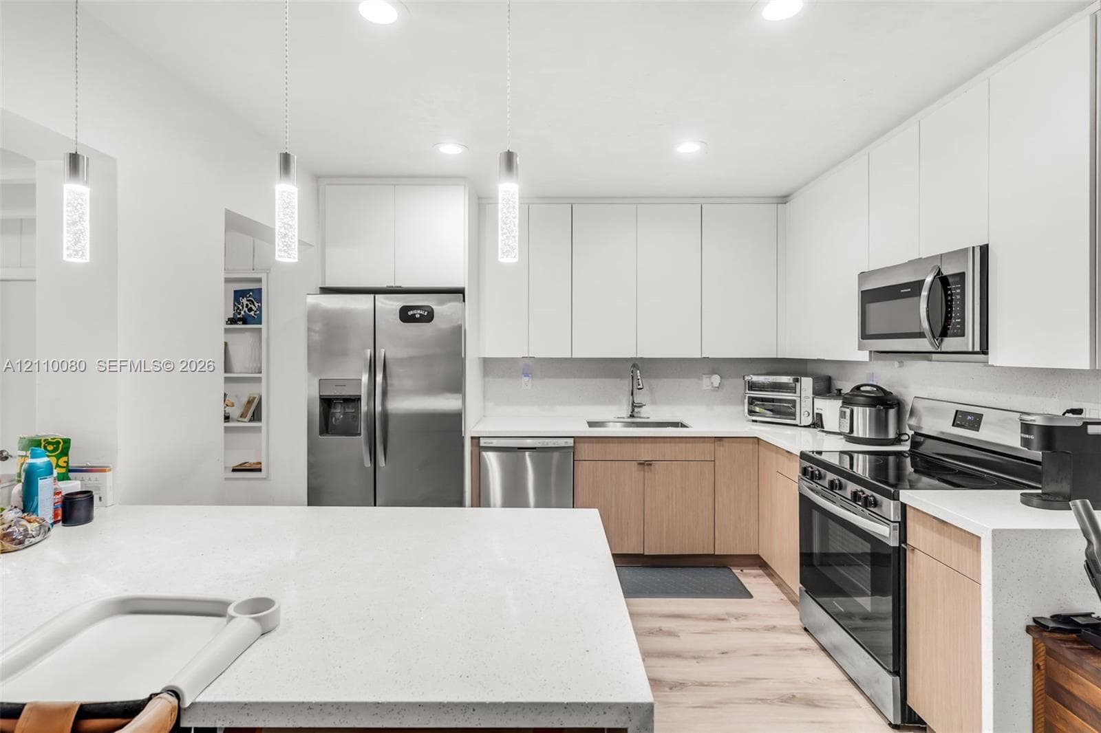 Kitchen featuring white upper cabinetry, light-toned wood-finish lower cabinetry, stone-composite countertops, stainless steel appliances, and minimalist pendant lighting