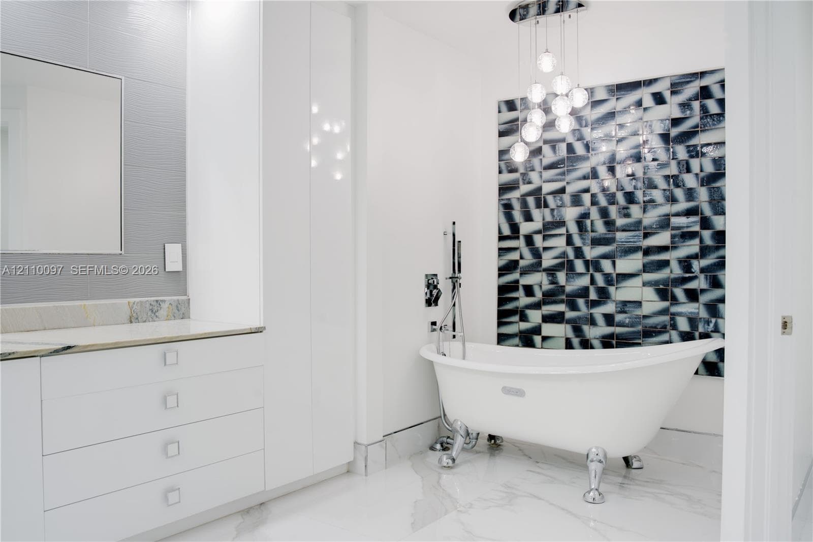 Clawfoot soaking tub with polished chrome hardware, geometric patterned accent wall, white cabinetry with integrated countertop, multi-globe pendant lighting, and large-format tile flooring