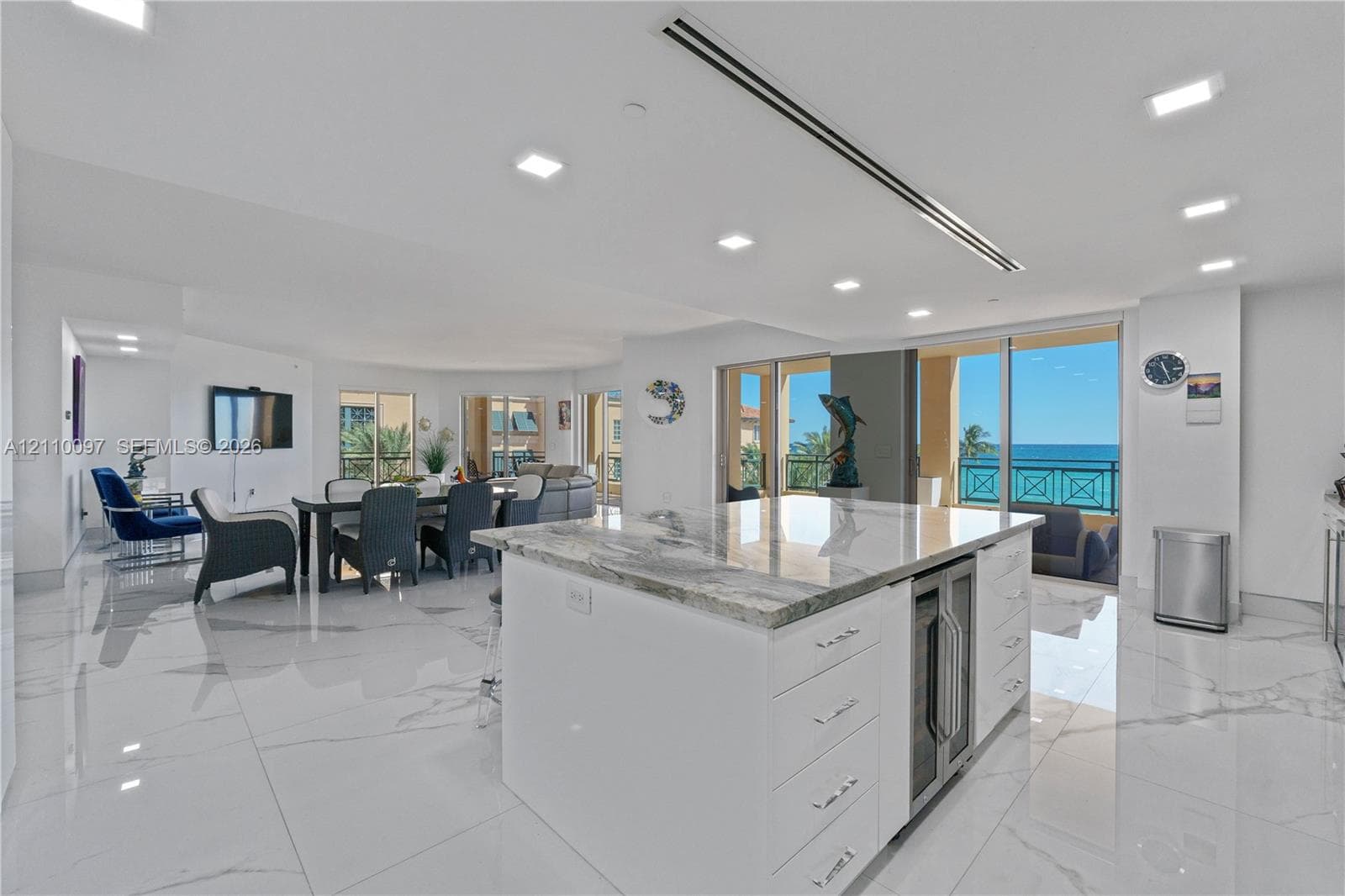 Open-concept kitchen featuring an expansive stone-topped island with an under-counter beverage chiller, high-gloss large-format tile flooring, recessed lighting, and expansive floor-to-ceiling glass doors opening to an exterior balcony