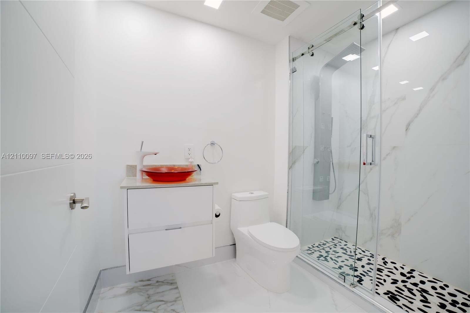 Floating white vanity with a colorful vessel sink, wall-mounted faucet, and walk-in shower featuring a glass sliding door, stone-patterned wall panels, and patterned floor tile