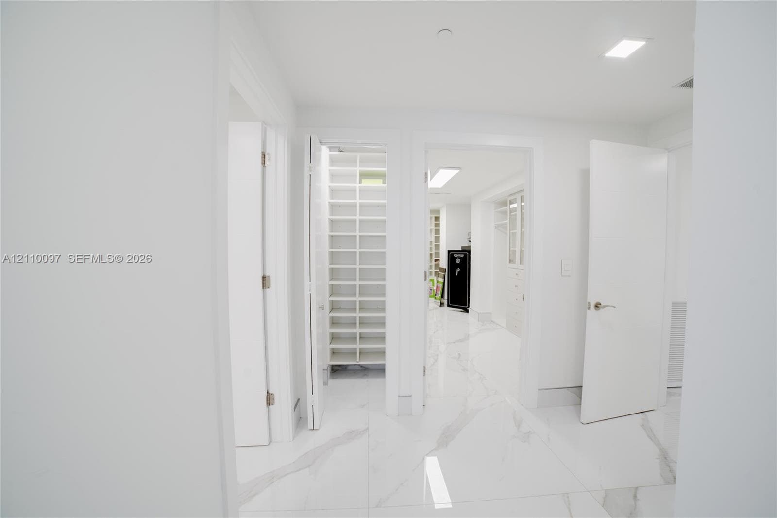 Large-format stone-finish flooring with white and grey veining throughout, featuring multiple walk-in storage areas with custom built-in shelving, white slab-style doors, and recessed lighting