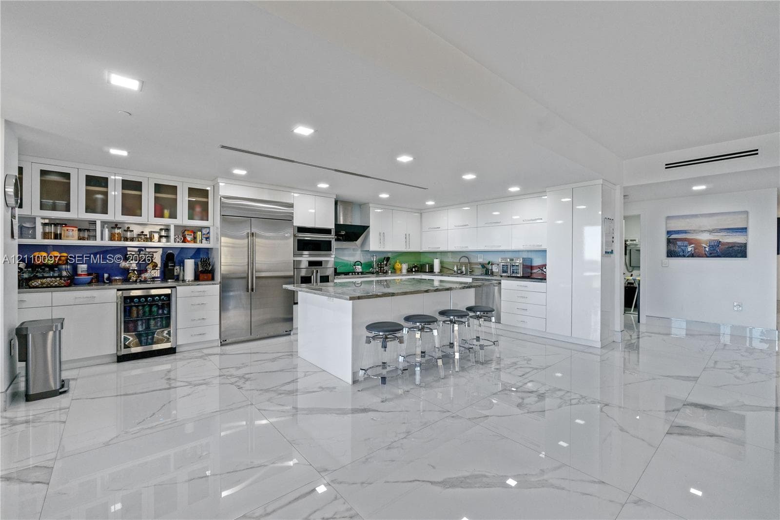 Modern kitchen featuring high-gloss white cabinetry, expansive stone-topped center island, large-format polished tile flooring, stainless steel appliances, and integrated wine refrigeration