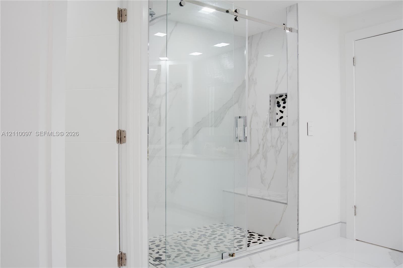 Walk-in shower featuring a frameless glass door, stone-finish wall tile with veining, pebble-style shower floor, built-in tiled bench, and an integrated recessed storage niche