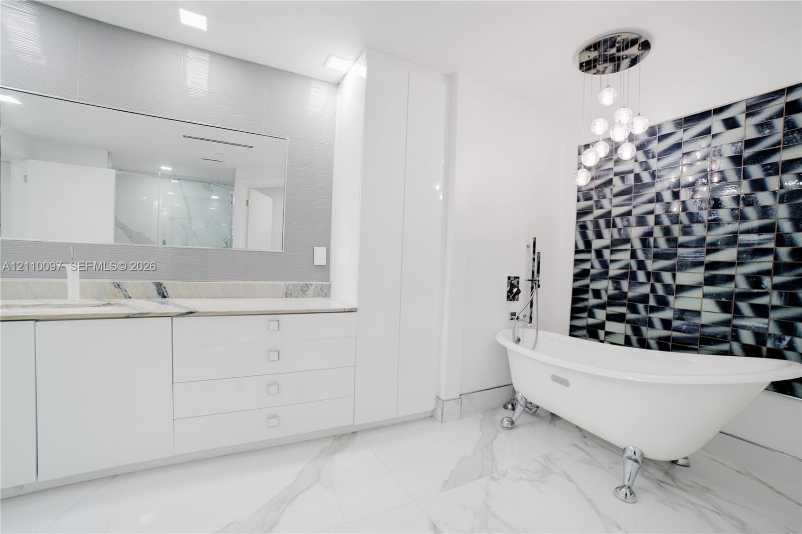 Freestanding clawfoot tub with chrome hardware, floor-to-ceiling geometric decorative wall tiling, custom white cabinetry with stone countertop, large-format veined flooring, and modern multi-globe chandelier lighting
