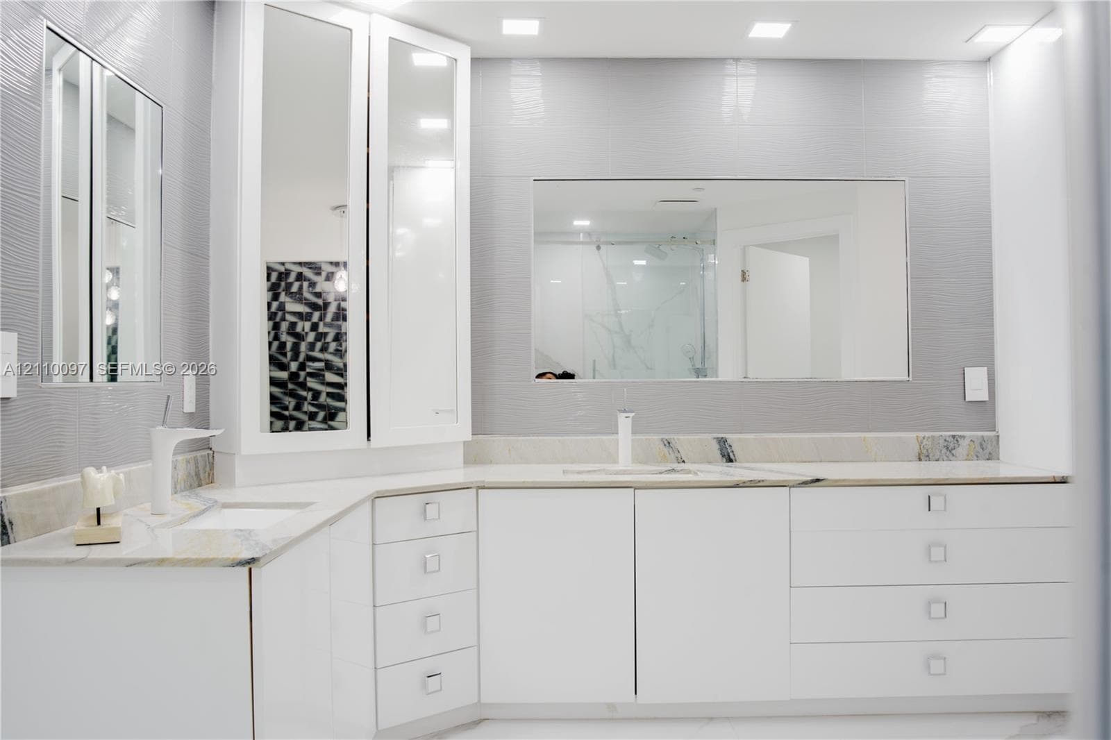 Custom white cabinetry with rectangular hardware, expansive countertop, dual recessed mirrors, and grey textured wall tiling