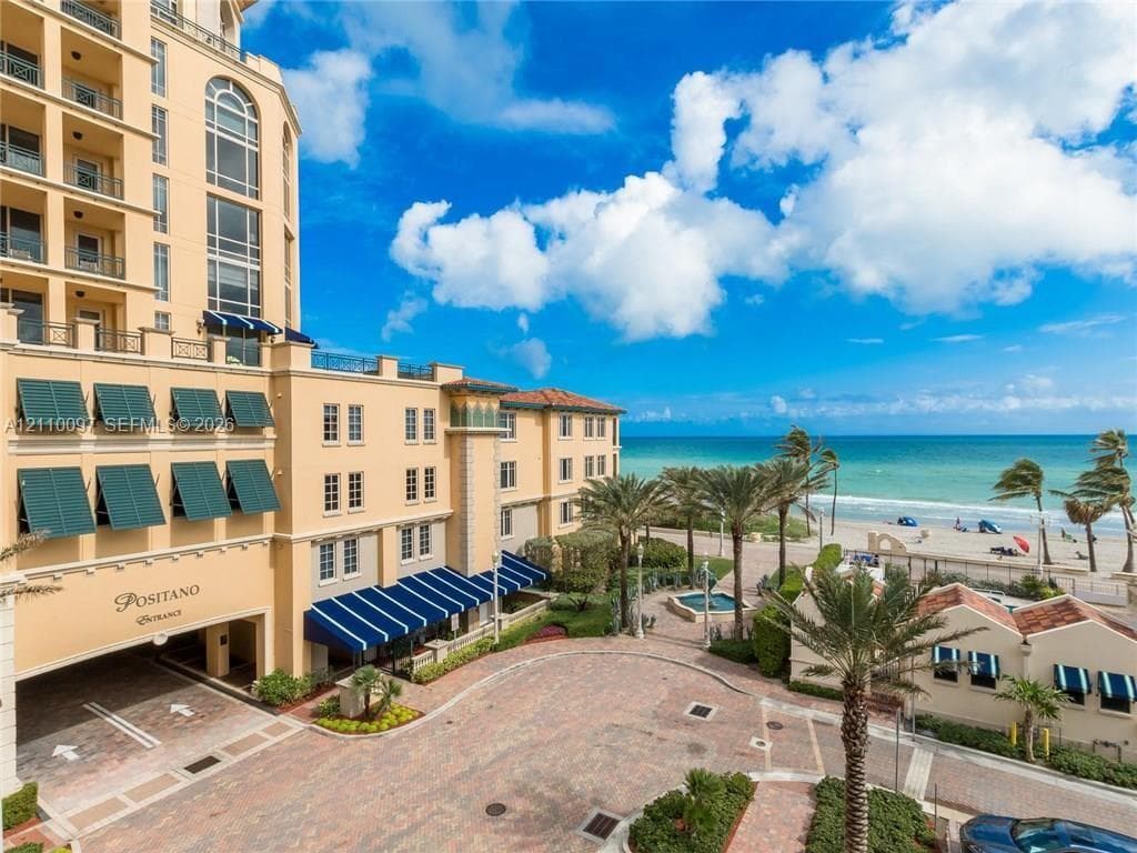 Multi-story Mediterranean-style exterior featuring a covered porte-cochère, blue fabric awnings, paver-stone driveway, and immediate proximity to the coastline