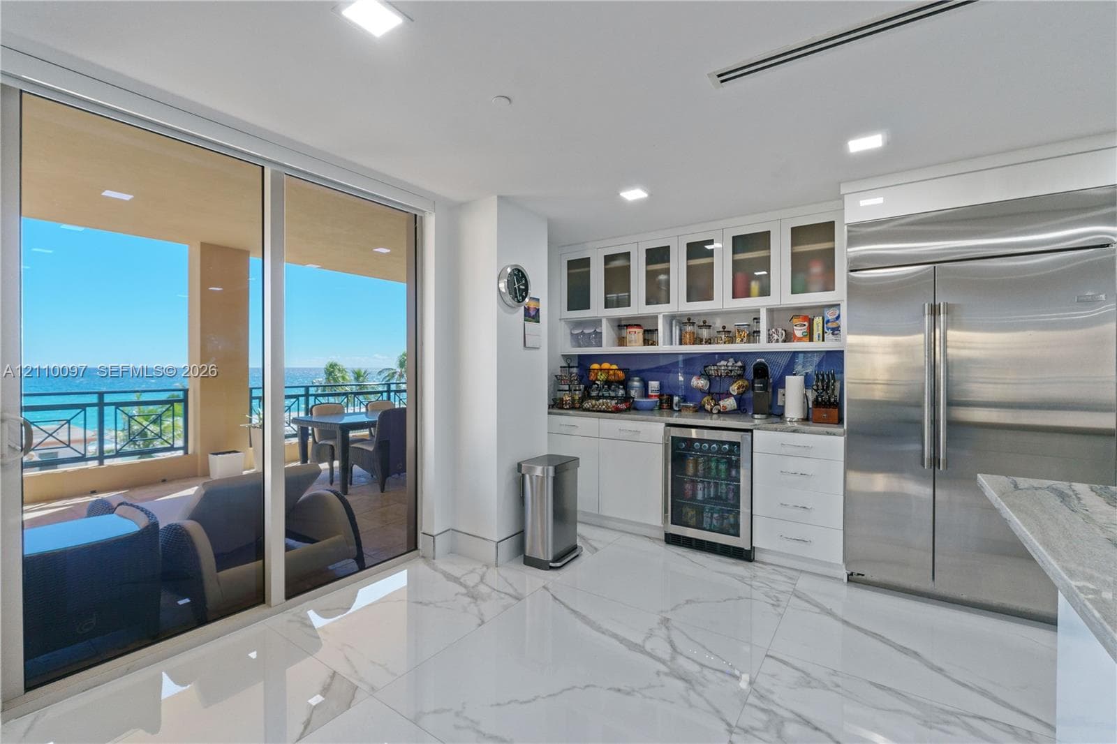 Large-format tile flooring, built-in white cabinetry with glass-front uppers, stainless steel side-by-side refrigerator, under-counter beverage cooler, and expansive floor-to-ceiling sliding glass doors opening to a covered balcony