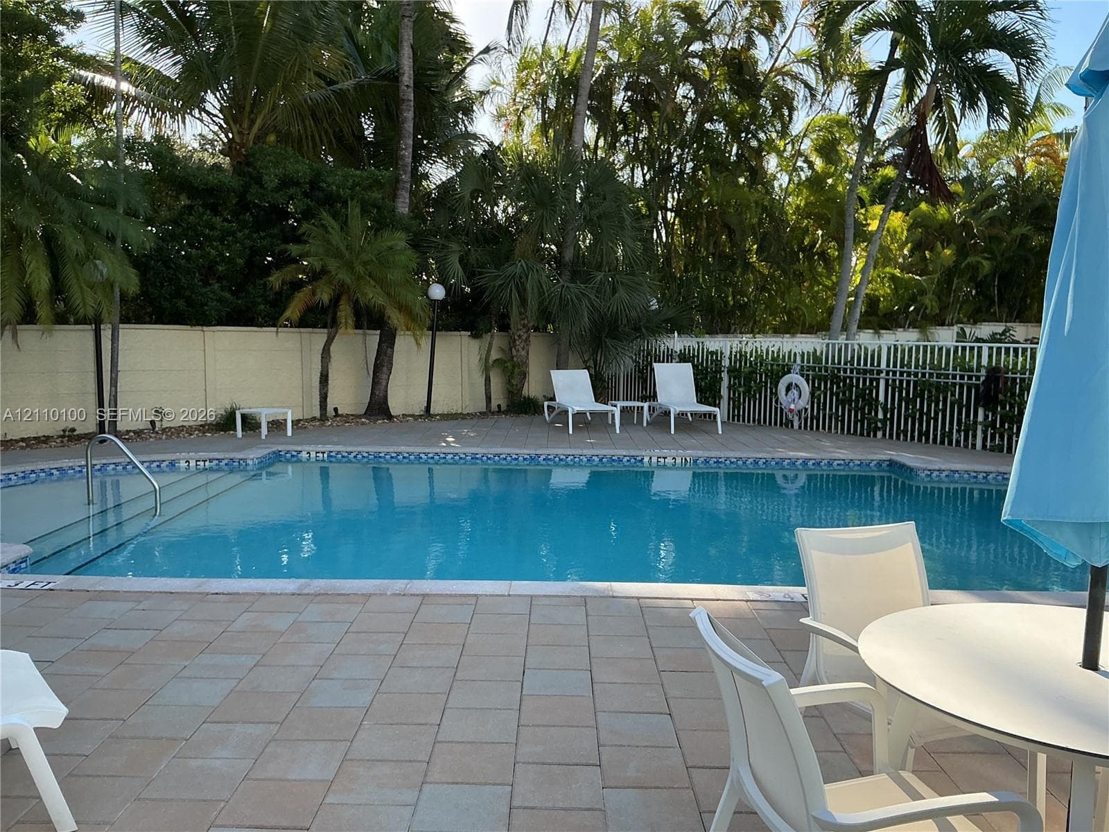 In-ground swimming pool with built-in stairs, surrounded by a paved patio, perimeter fencing, and mature palm trees