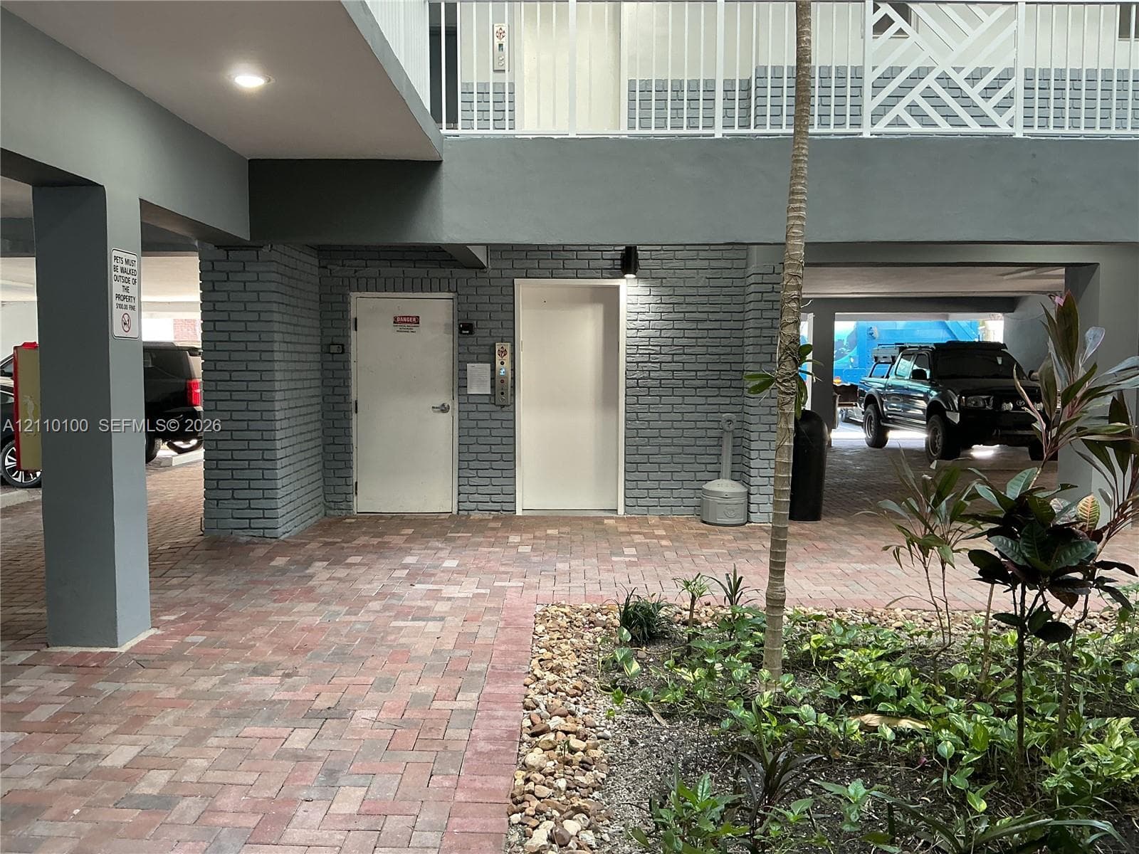 Covered ground-level parking area featuring paver flooring, painted brick structural columns, an elevator, and landscaped garden beds