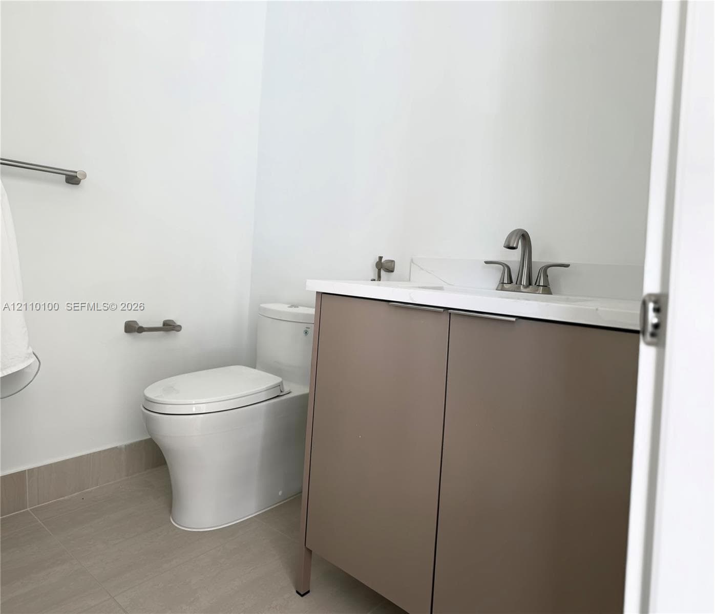 Modern vanity with a neutral-toned cabinet, white stone-composite countertop, dual-handle metal faucet, and floor-mounted commode, complemented by tile flooring