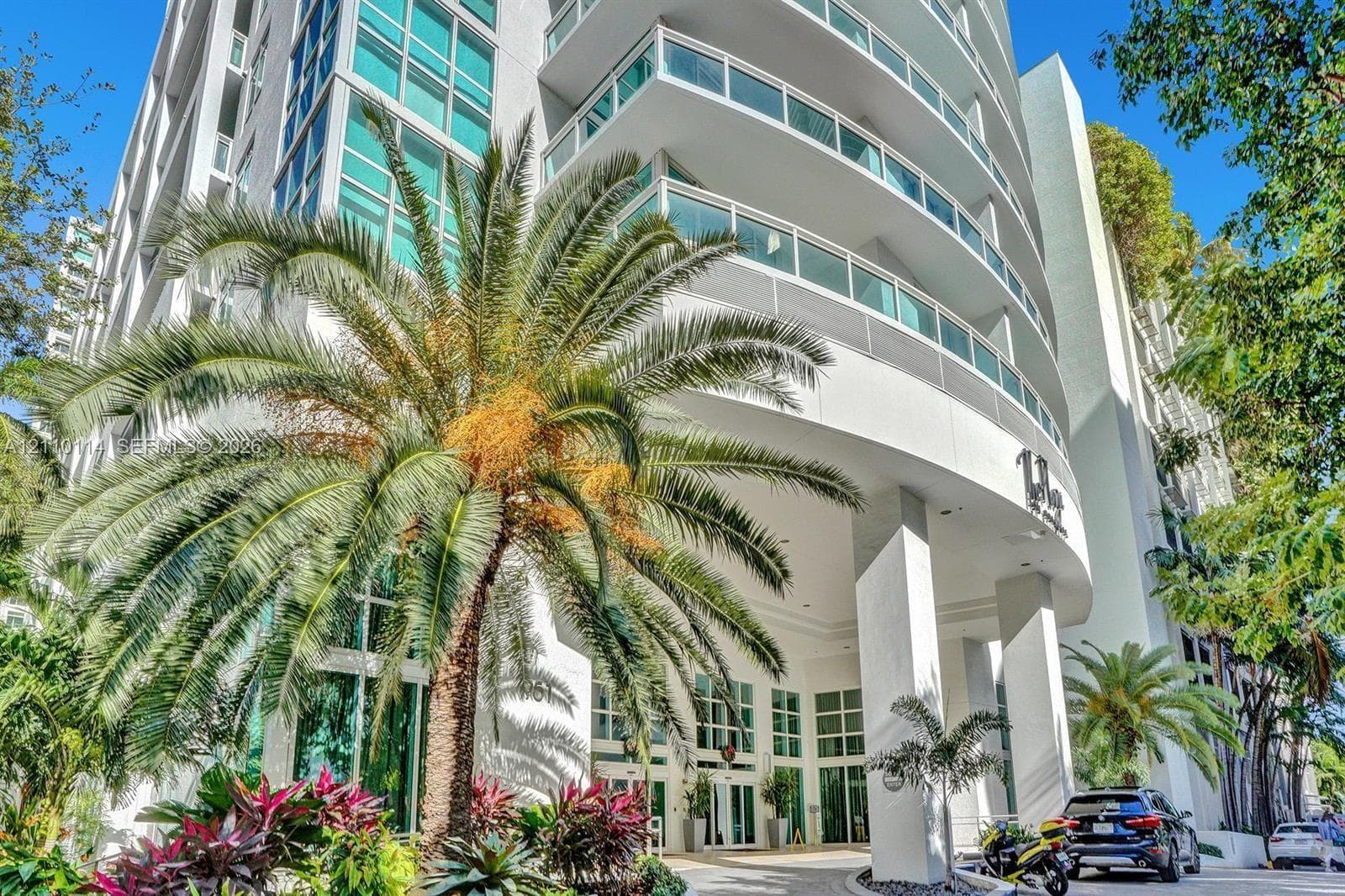 Modern high-rise exterior featuring curved balconies, floor-to-ceiling glass windows, concrete structure, and ground-level landscaping