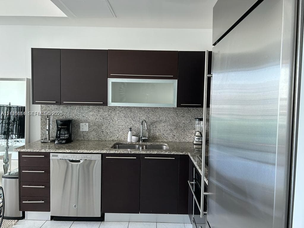 Kitchen featuring dark-finish cabinetry, stone countertop, stainless steel dishwasher, integrated stainless steel refrigerator, and undermount dual-basin sink with metallic faucet