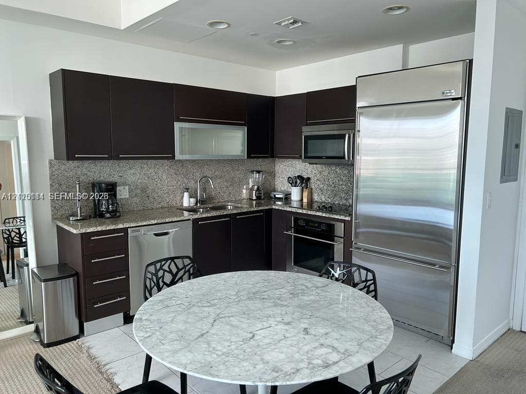 Kitchen featuring dark-toned cabinetry, light stone countertops, integrated stainless steel refrigerator, built-in oven, and recessed lighting