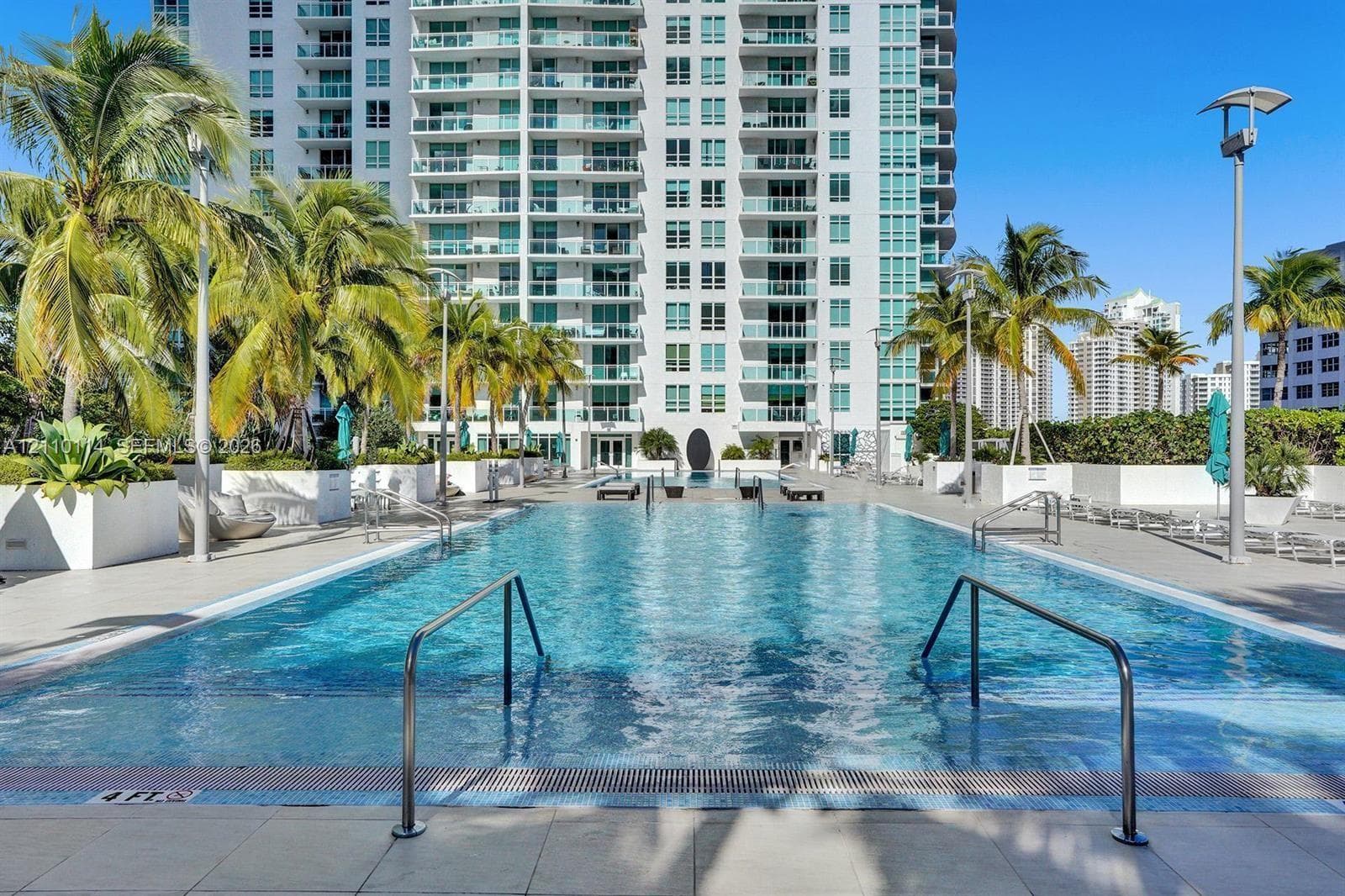 Rectangular swimming pool with integrated entry steps and stainless steel handrails, surrounded by a paved deck, large white planters, and mature palm trees