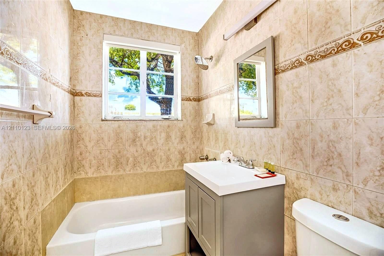 Full-height wall tiling with decorative border, bathtub-shower combination, grey vanity cabinet with integrated white sink, rectangular mirror, and bar lighting
