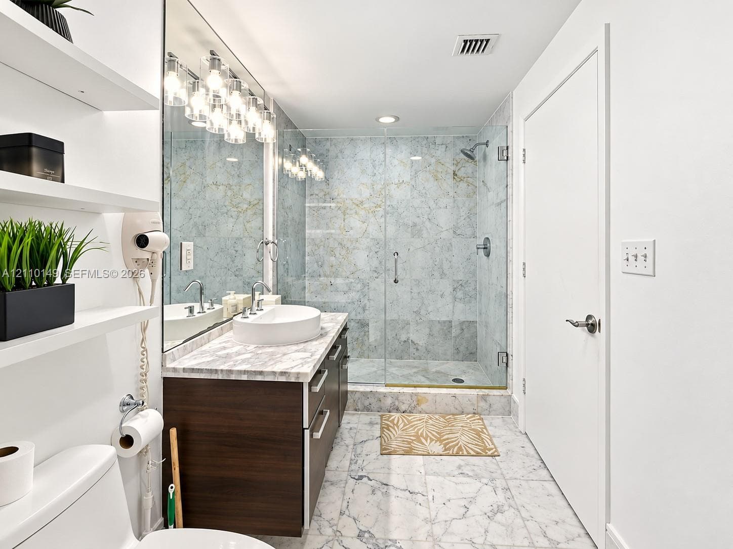 Bathroom featuring a dark wood-finish vanity with a stone-top surface and vessel sink, walk-in shower with floor-to-ceiling tile surround and glass enclosure, wall-to-wall mirror, built-in shelving, and light-toned tile flooring