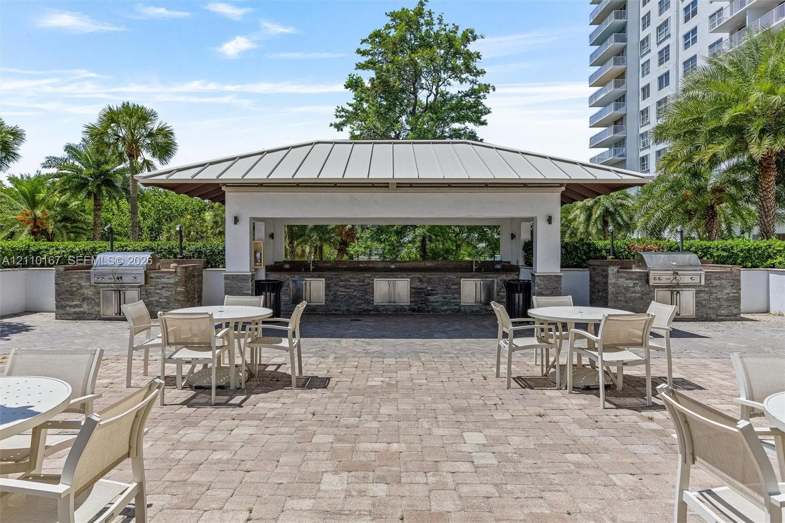 Outdoor pavilion featuring a stone-clad kitchen island, dual built-in stainless steel grills, a metal-seam roof, and extensive stone paver patio space