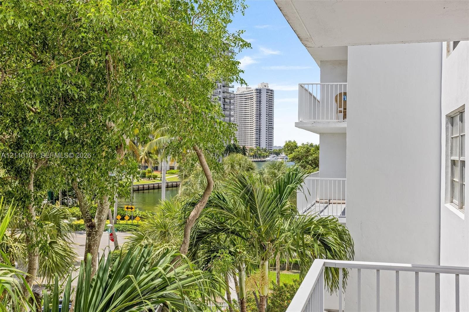Private balcony with white metal railings, overlooking lush landscaping, mature trees, and a canal