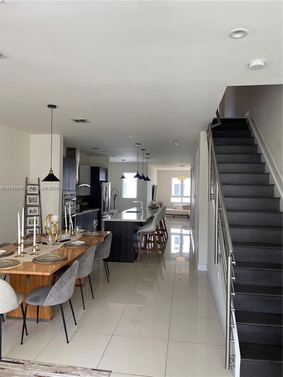 Open-concept interior featuring large-format tile flooring, a kitchen island with stone countertop, dark-toned cabinetry, recessed lighting, and a staircase with metal railings