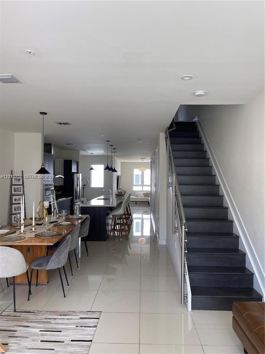 Open-concept interior featuring large-format tile flooring, a kitchen island with stone countertop, stainless steel pendant lighting, and a staircase with metal railings