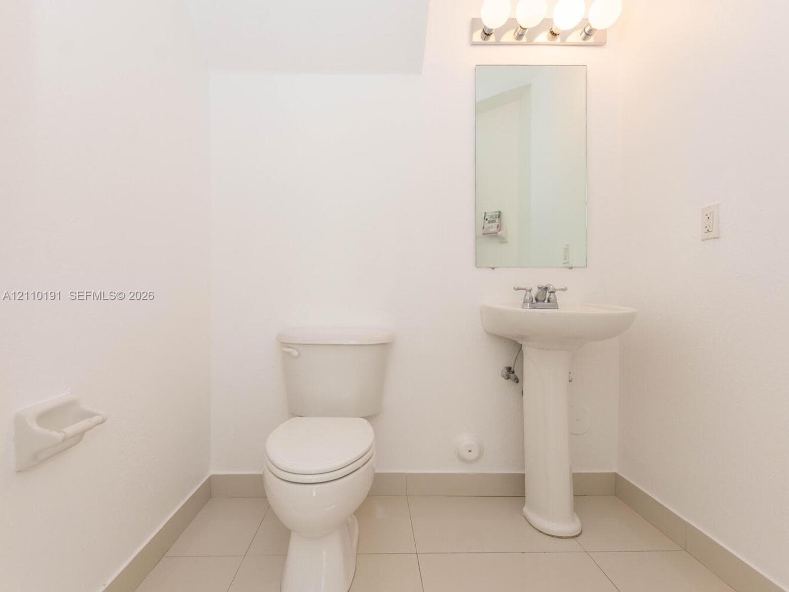 Bathroom featuring a white pedestal sink, dual-handle faucet, wall-mounted rectangular mirror, four-light vanity fixture, and tiled flooring