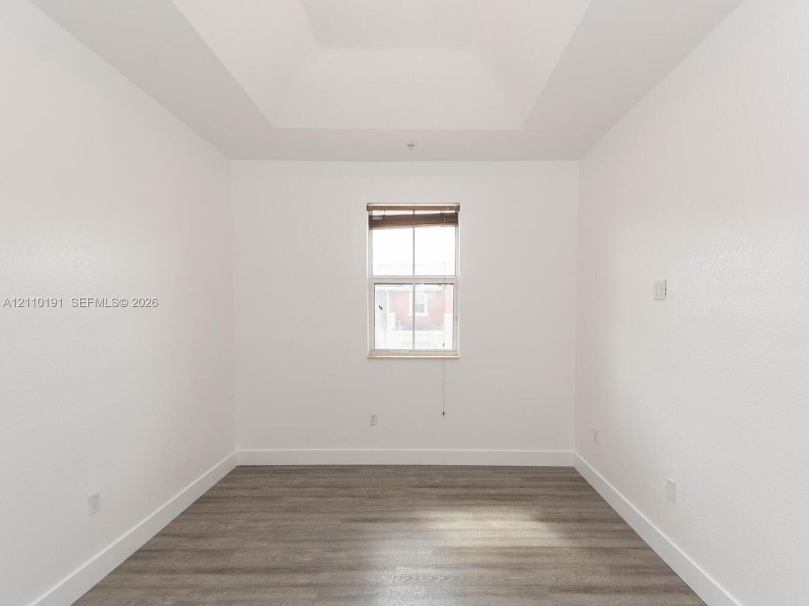 Room featuring tray ceilings, wood-finish flooring, and a central window with integrated blinds