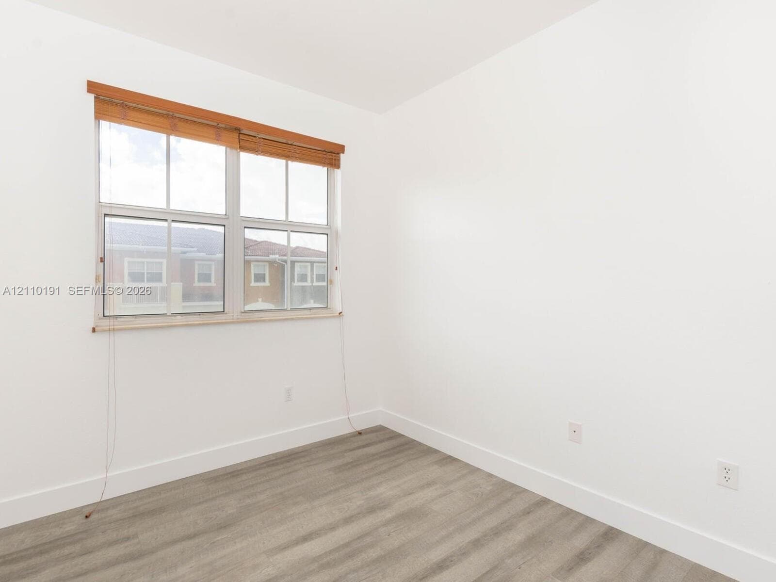 Neutral-toned interior space featuring wood-finish flooring, crisp white walls, and a multi-pane window with wood-slat blinds