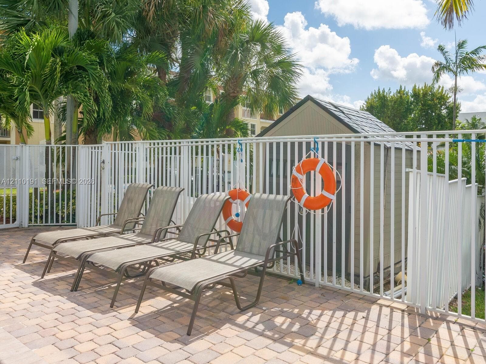 Paver patio featuring a perimeter metal fence, a utility shed, and mature palm trees