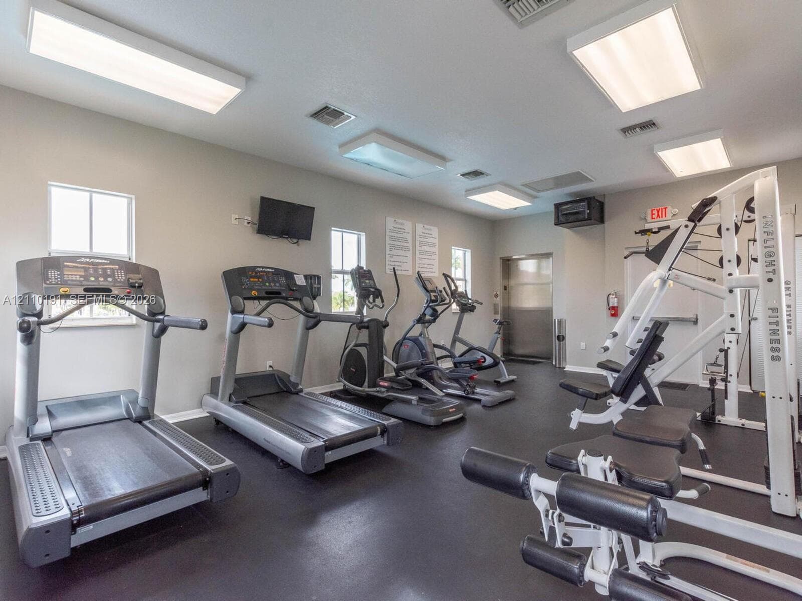 Fitness center featuring rubber flooring, multiple rectangular ceiling-mounted light fixtures, and windows providing natural light